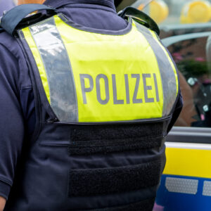 German police officer seen from behind in a yellow vest with Polizei text, standing by patrol car. Suitable as background for  breaking news, reports, or public safety and law enforcement topics.