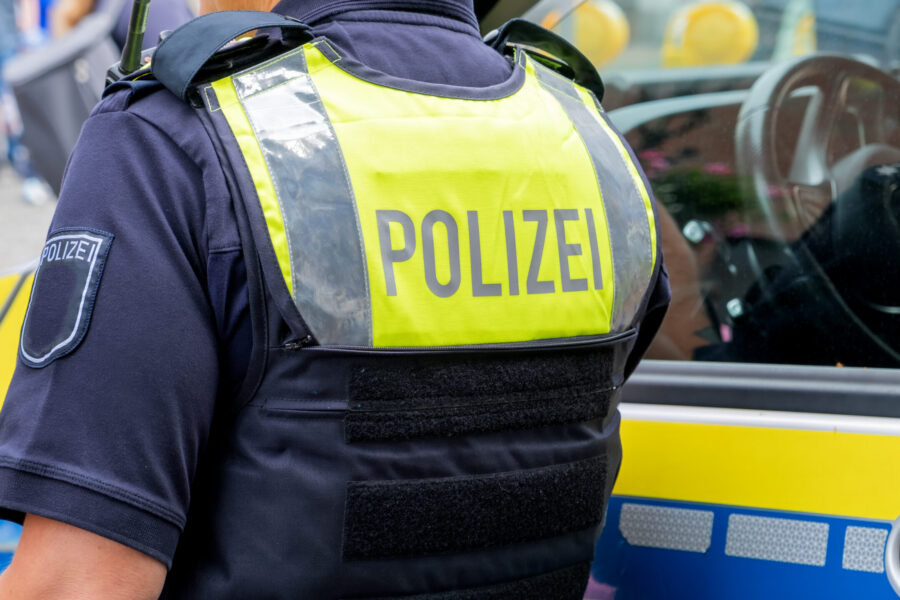 German police officer seen from behind in a yellow vest with Polizei text, standing by patrol car. Suitable as background for  breaking news, reports, or public safety and law enforcement topics.